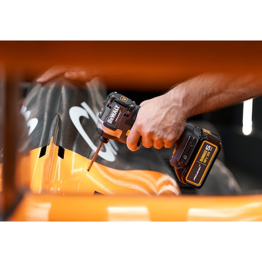 A close-up of a person's hand using a DEWALT cordless impact driver on a carbon fiber orange and black surface, possibly