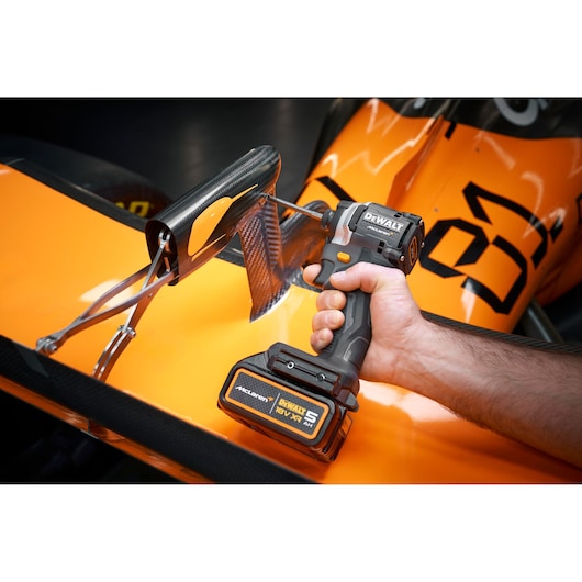 A close-up of a person using a DEWALT cordless drill on a part of an orange race car, working on a carbon fiber component.