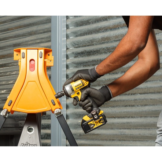 A person wearing black gloves is using a DEWALT cordless impact wrench (DCF880M2-QW) to tighten screws on an orange metal structure in an industrial setting.
