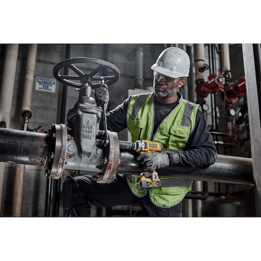 A worker wearing a green safety vest and helmet is using a DEWALT DCF891P2T power tool to work on a large industrial valve attached to metal pipes inside a facility.