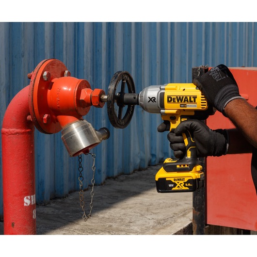 A person wearing gloves is using a DEWALT cordless impact wrench (model DCF899P2-QW) to adjust a valve on a red industrial pipe near a blue metal wall.