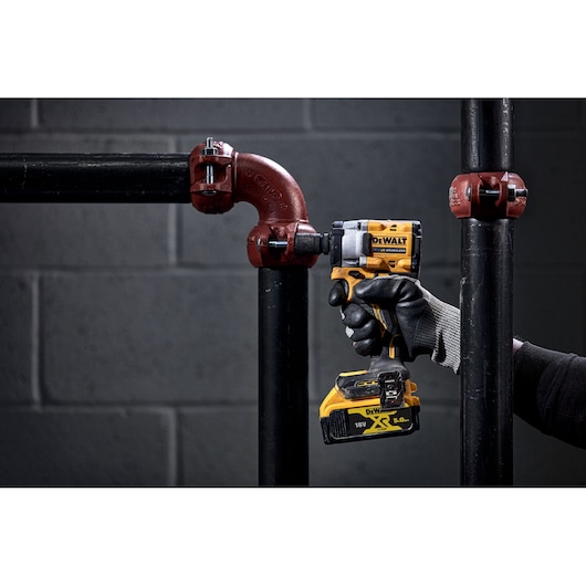 A person wearing gloves uses a DEWALT cordless impact wrench to fasten a bolt on a metal pipe fitting. The tool is yellow and black, and the scene is set against a dark, industrial background.