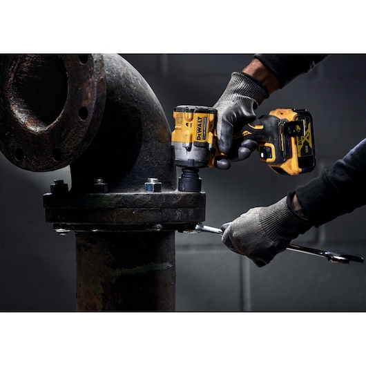 A person wearing work gloves uses a DEWALT DCF922P2T cordless impact wrench to tighten bolts on a large industrial pipe joint.