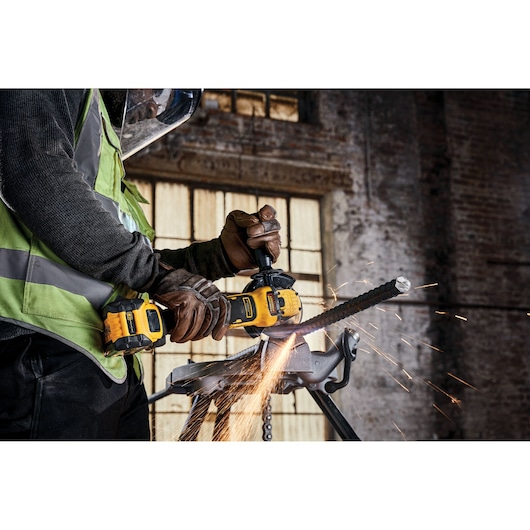 A close-up of a person using a yellow and black DEWALT cordless angle grinder to cut through a metal rebar, with sparks flying. The person is wearing work gloves, a safety vest, and a face shield, and is working in an industrial setting.
