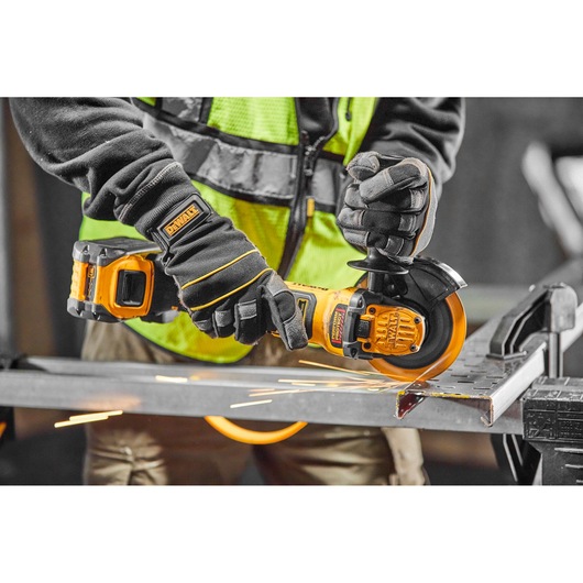 A person wearing protective gloves and a high visibility vest uses a DEWALT cordless angle grinder to cut a metal piece, with visible sparks flying.