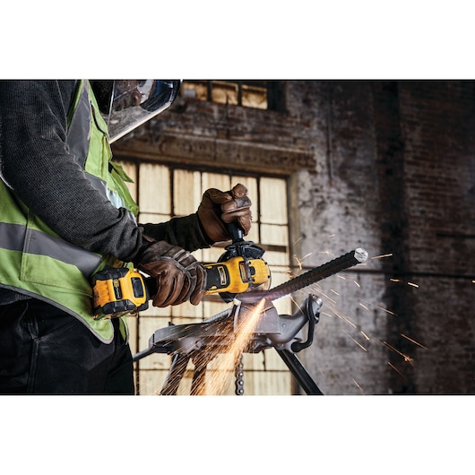 A person wearing protective gear uses a DEWALT cordless angle grinder (model DCG409VSN) to cut a metal rebar, creating sparks in an industrial workshop setting.