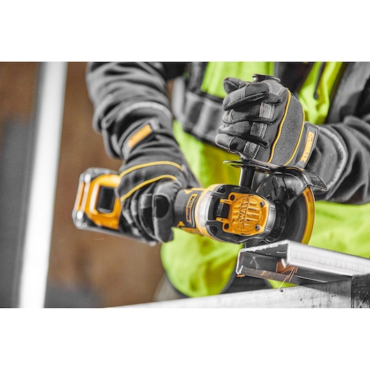A close-up of a person wearing gloves and using a DEWALT cordless angle grinder to cut a metal piece. The person is dressed in a high-visibility vest and dark clothing.