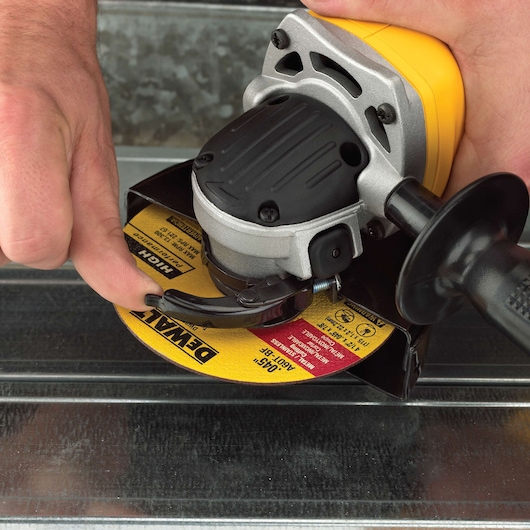 Close-up of a person adjusting a DEWALT angle grinder. The hand is turning a lever near the yellow DEWALT cutting disc, with the tool placed on a metallic surface.