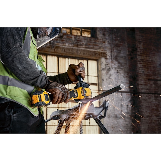 A person wearing safety gear and gloves uses a DEWALT cordless angle grinder to cut a metal rod, with sparks flying, inside