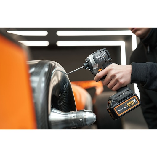 A person uses a DEWALT cordless impact driver to fasten a screw on a carbon fiber component, possibly part of a vehicle, in a modern industrial setting.