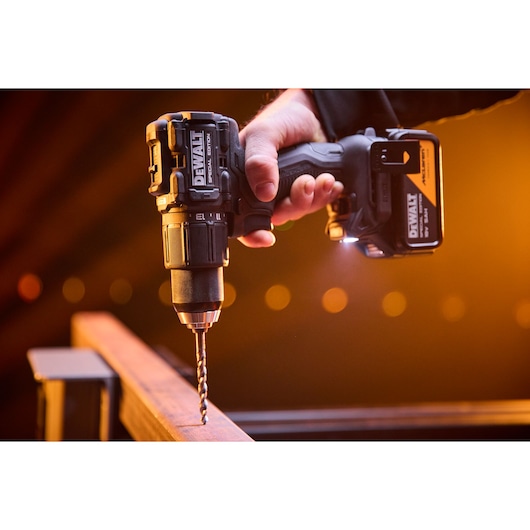 A DEWALT cordless drill in use, drilling into a metal beam with warm lighting in the background.