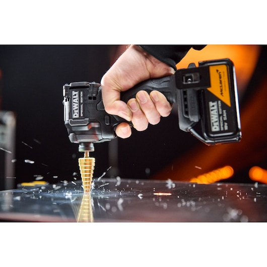 Hand uses DEWALT cordless drill with step bit to drill through metal sheet, sparks flying.