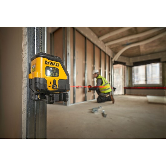 The DEWALT USB Red Beam Cross Line Laser in foreground attached to metal frame using the magnetic wall bracket, red laser is on and worker is onsite in the background.