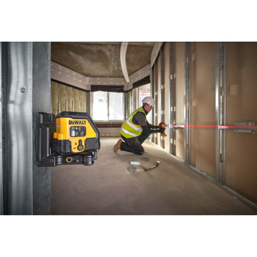 A DEWALT laser level is mounted on a metal frame inside a building under construction. In the background, a person wearing a