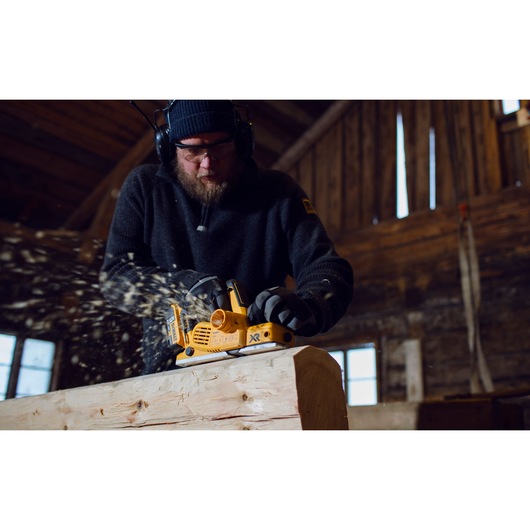 A person using a DEWALT DCP580P2 cordless planer to smooth a piece of wood inside a workshop, with wood shavings flying. The person's face is obscured and they are wearing protective headphones and gloves.
