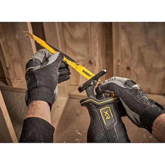 Gloved hands holding DEWALT reciprocating saw, inserting a yellow blade in a woodwork setting.