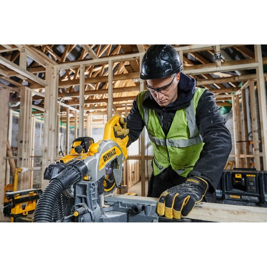 A person wearing protective gear and a high-visibility vest is using a DEWALT miter saw to cut a wooden plank on a construction site with exposed wooden framing.