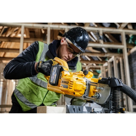 A person wearing safety gear operates a DEWALT DCS777T2 power tool, cutting wood in a construction site. The tool is bright yellow, and the person is in a partially built wooden structure.