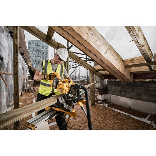 A construction worker wearing a safety helmet and high-visibility vest uses a DEWALT DCS777T2 cordless miter saw to cut a wooden beam at a building site. The saw is mounted on a stand, and the work area is surrounded by exposed beams and partially built walls.