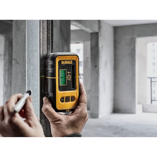A person is holding a DEWALT DE0892G green beam laser detector against a metal beam in a construction site, using a marker to align with a laser line.