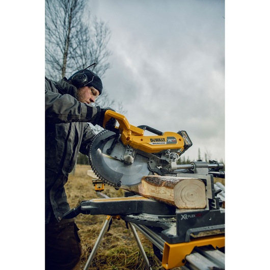 A person using a DEWALT DHS780T2A cordless mitre saw to cut a piece of wood outdoors. The saw is yellow and mounted on a