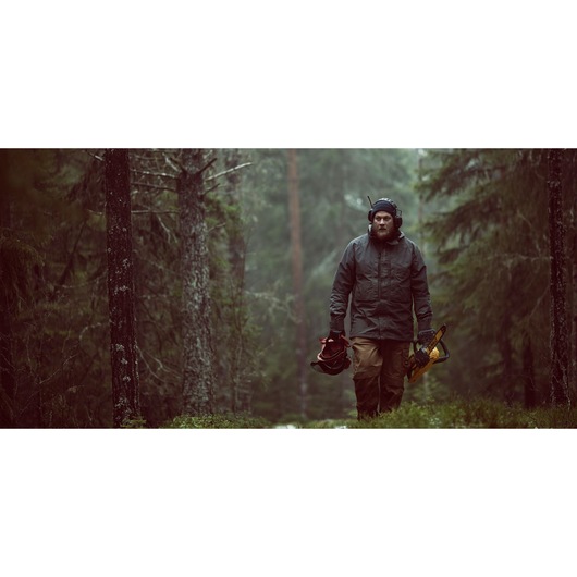 A person wearing protective gear walks through a forest while carrying a DEWALT chainsaw and a helmet.