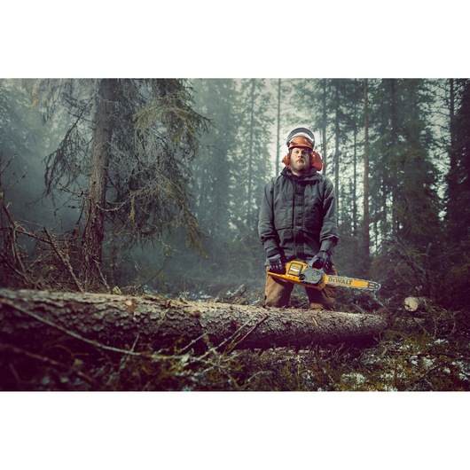 A person wearing safety gear stands in a misty forest, holding a yellow DEWALT chainsaw. There is a large felled tree in the
