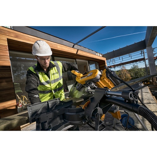 A person wearing a hard hat and high-visibility vest uses a DEWALT DHS780T2A miter saw to cut wood on an outdoor
