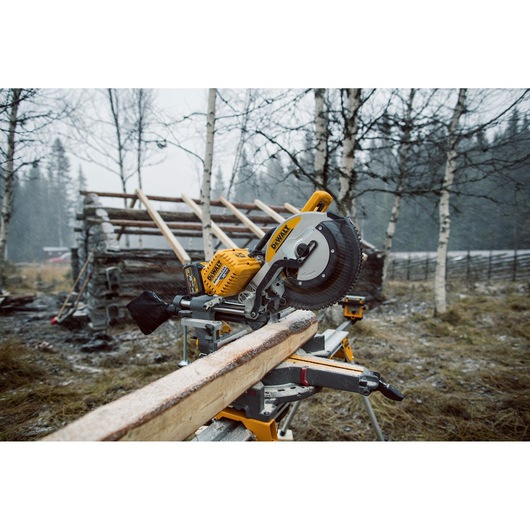 A DEWALT DHS780T2A yellow and black cordless mitre saw placed outdoors, cutting a wooden beam. The saw is set up in a
