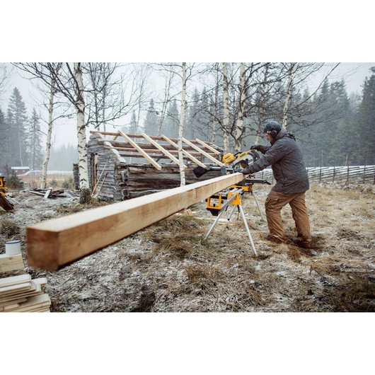 A person using a DEWALT miter saw on a worksite outdoors, cutting a long wooden beam. The scene is set in a snowy, rural