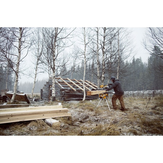 A person using a DEWALT miter saw in an outdoor setting, cutting a wooden beam next to a partially constructed log cabin