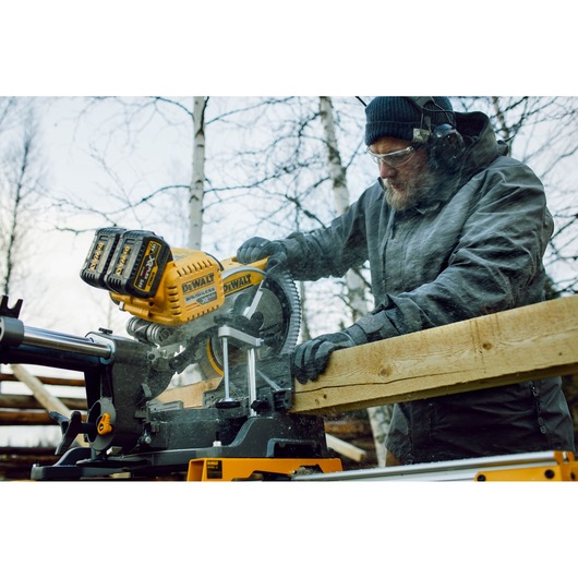 A person using a DEWALT DHS780T2A cordless mitre saw to cut a wooden plank outdoors. The operator is wearing protective