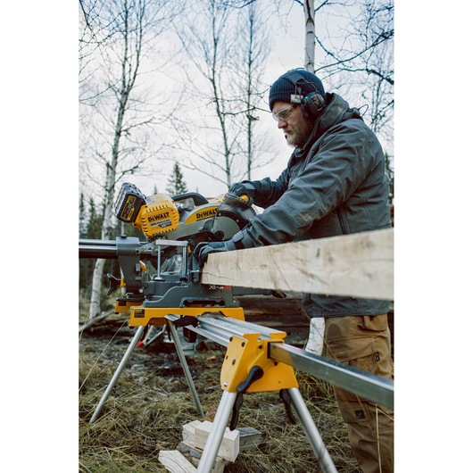 A person wearing protective earmuffs and gloves operates a DEWALT battery-powered miter saw, cutting a piece of wood