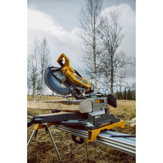 A DEWALT cordless mitre saw cutting a piece of wood outdoors, set up on a metal stand with bare trees and grass in the
