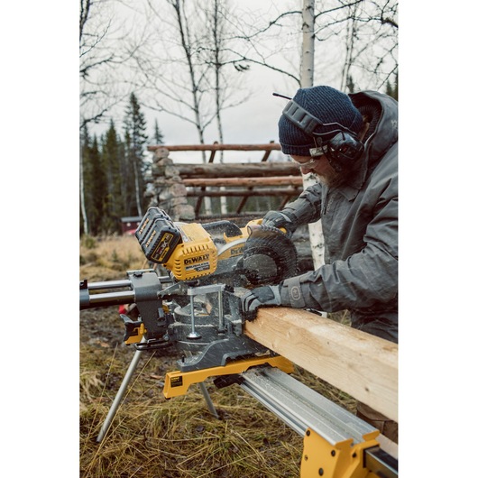 A person wearing protective gear uses a DEWALT DHS780T2A cordless miter saw to cut a wooden beam outdoors. The saw is