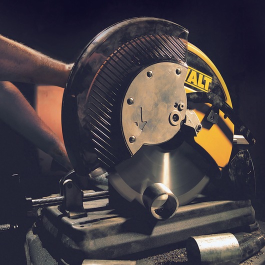 A DEWALT metal cutting saw in use, cutting through a cylindrical metal pipe. The saw is mounted on a base, and a person's