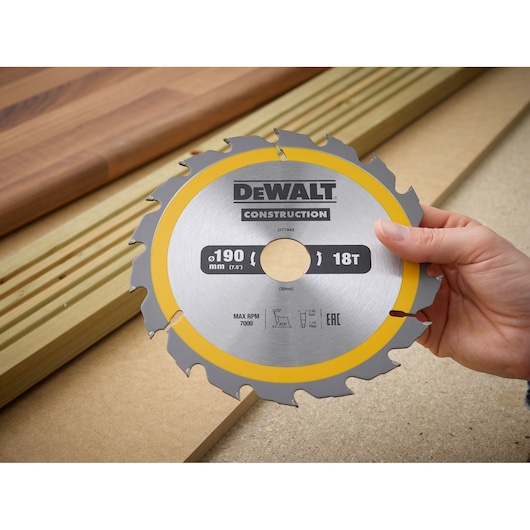 A hand holds a DEWALT DT1943 circular saw blade, 190mm 18T, with wood planks in the background.