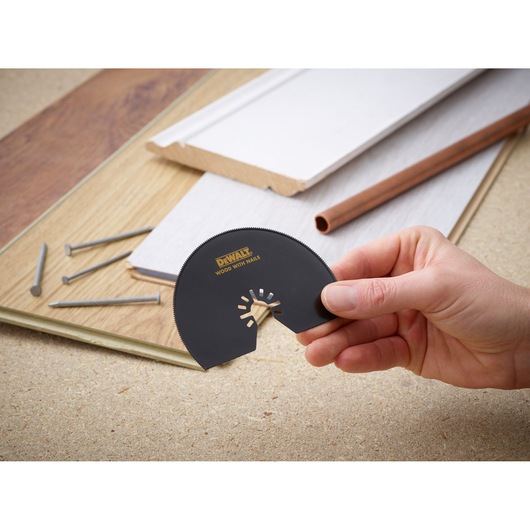 A hand holding a DEWALT DT20708 oscillating multi-tool blade, labeled 'WOOD WITH NAILS', with various construction materials, nails, and wooden planks in the background.