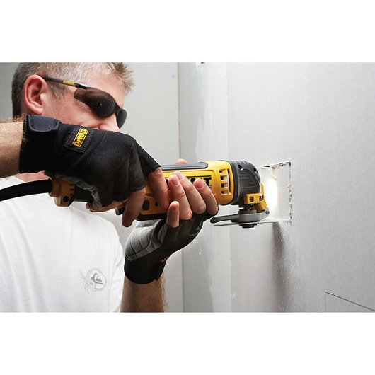 A person wearing protective gloves uses a yellow DEWALT oscillating multi-tool to cut a square opening in a drywall panel.