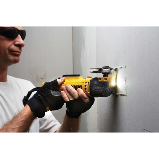 A person wearing gloves uses a DEWALT oscillating multi-tool to cut a rectangular hole into a drywall panel. The tool is