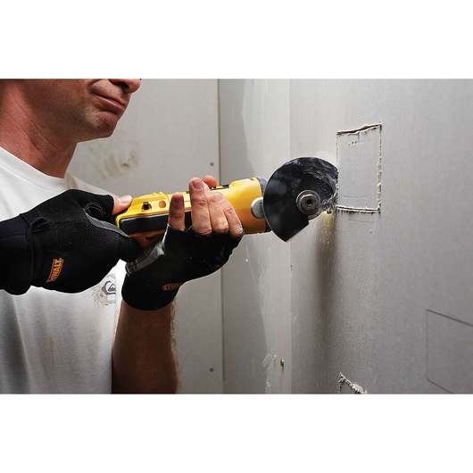 A person wearing black DEWALT gloves is using a yellow DEWALT power tool with a cutting blade to cut a rectangular hole in a