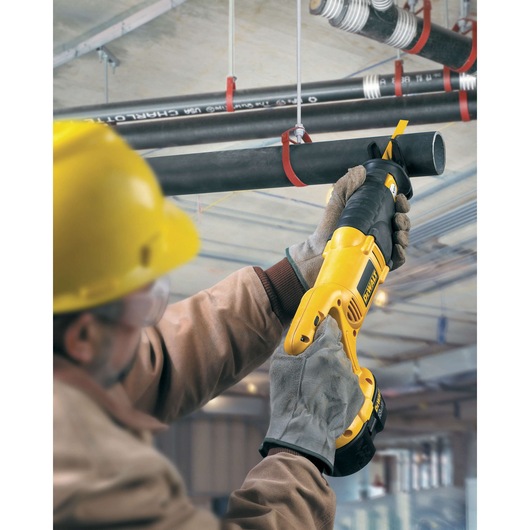 A worker wearing safety gear uses a DEWALT reciprocating saw to cut through a black pipe suspended from the ceiling in an