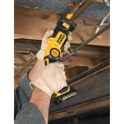 A person wearing gloves uses a DEWALT cordless power tool to cut a metal pipe in a workshop or construction area.
