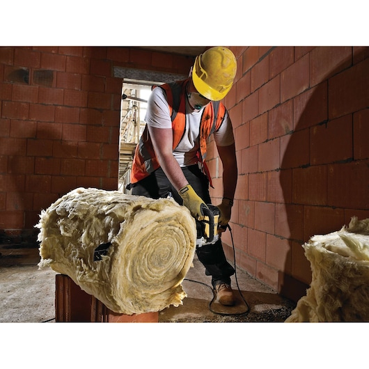A construction worker wearing a yellow hard hat, orange safety vest, gloves, and boots is using a DEWALT power tool to cut a