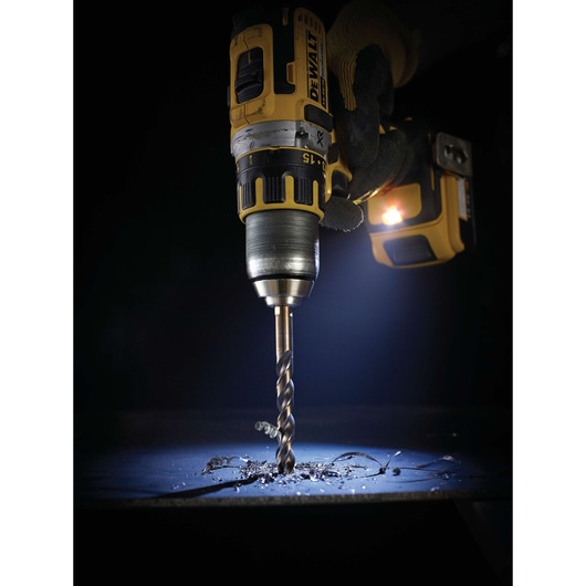 Close-up of a DEWALT cordless drill held by a gloved hand, drilling into a metal surface with metal shavings scattered
