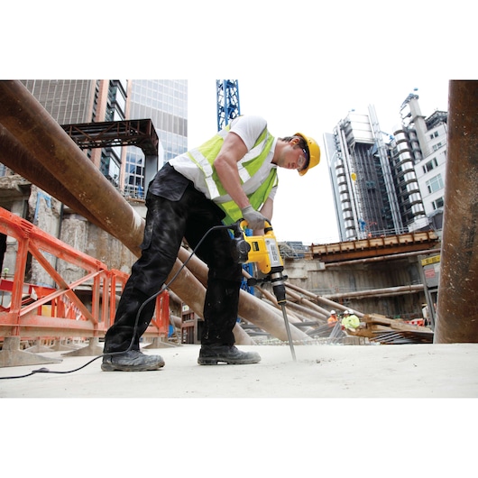 A construction worker wearing a yellow hard hat and reflective vest is using a DEWALT rotary hammer drill (SKU: DT6824) to