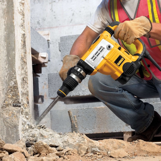 A person wearing gloves and a safety vest operates a yellow DEWALT rotary hammer drill, breaking into a concrete surface at
