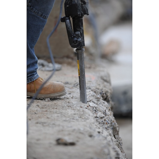A worker uses a DEWALT demolition tool to break concrete. Only the tool and boots are visible.