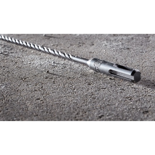 A close-up image of a DEWALT DT8914 SDS-plus drill bit lying on a rough concrete surface. The metal bit features a spiral design and is designed for masonry and concrete drilling.