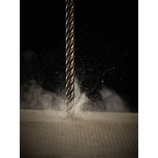 Close-up of a metal drill bit drilling into a concrete surface, with dust and debris flying up.
